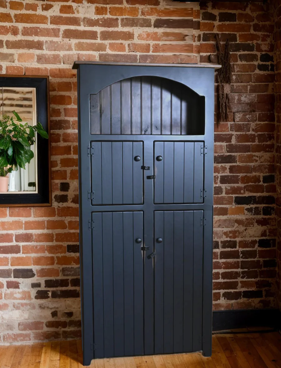 "Handcrafted American-made rustic storage cabinet with a distressed black finish, arched open shelf, and four-door design, set against an exposed brick wall."