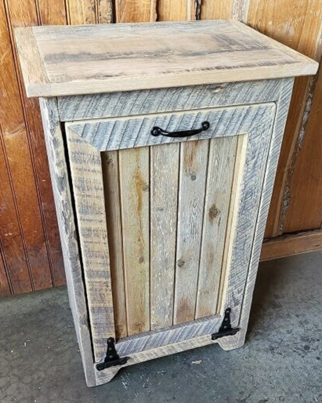 Rustic wooden tilt-out trash bin, handmade in the USA with black hardware and natural distressed wood, placed against a wooden wall.