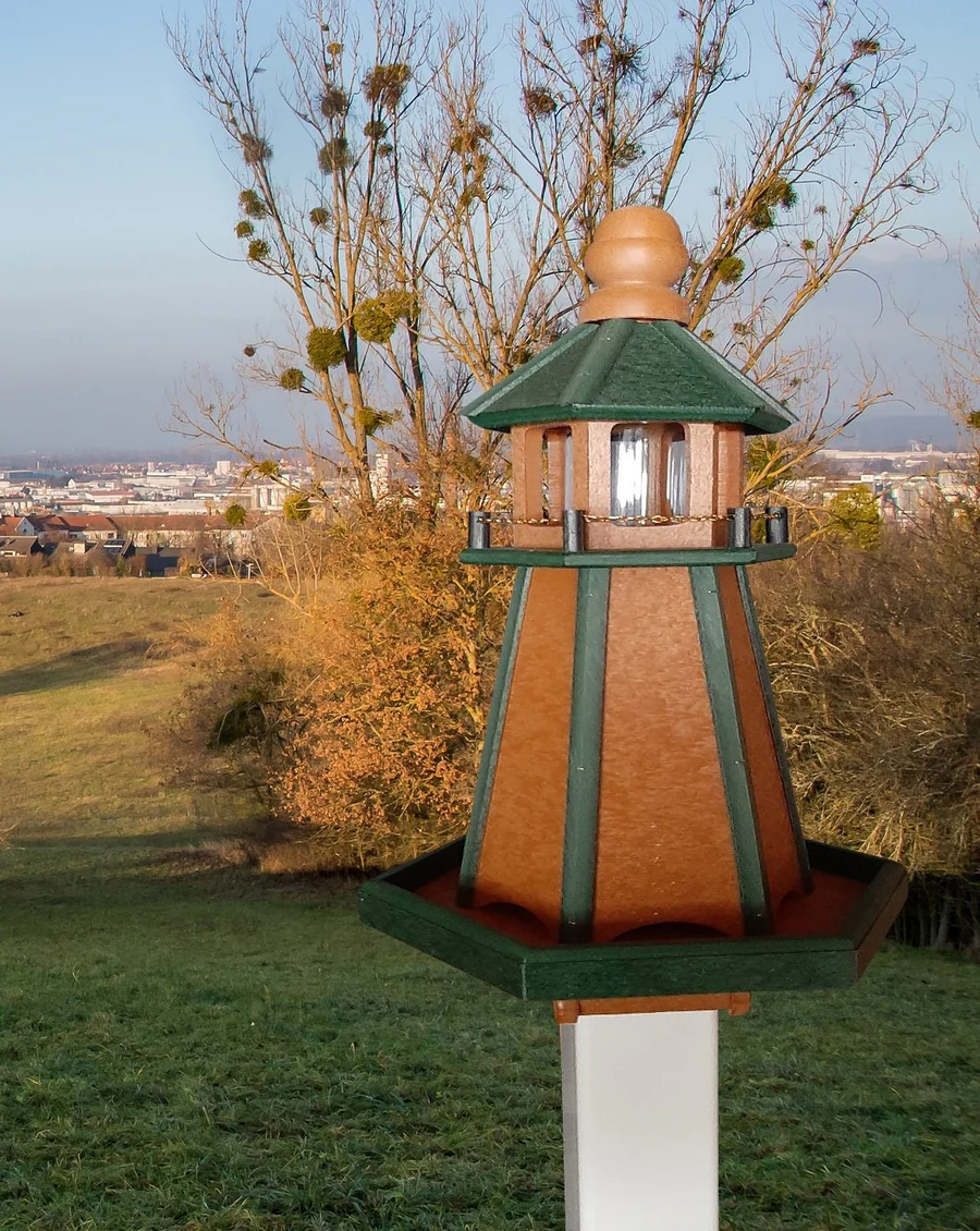 Wooden bird feeder with lighthouse-inspired design in green and tan, mounted on a white post with scenic background