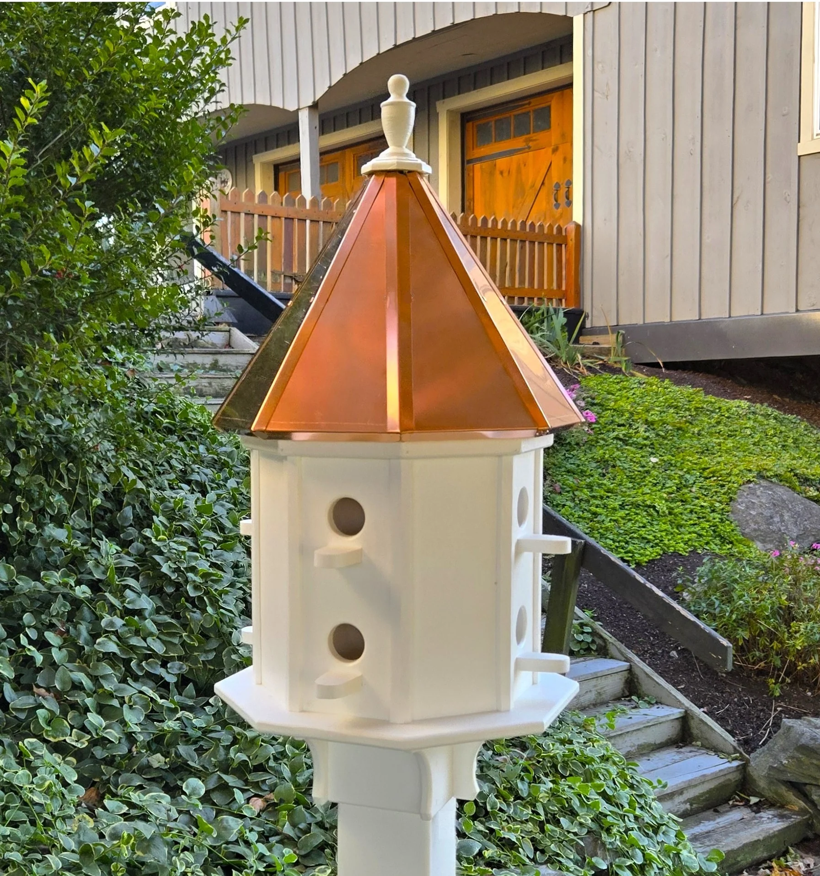 Elegant multi-compartment birdhouse with a copper roof and white post, surrounded by greenery and steps, adding charm to an outdoor setting.