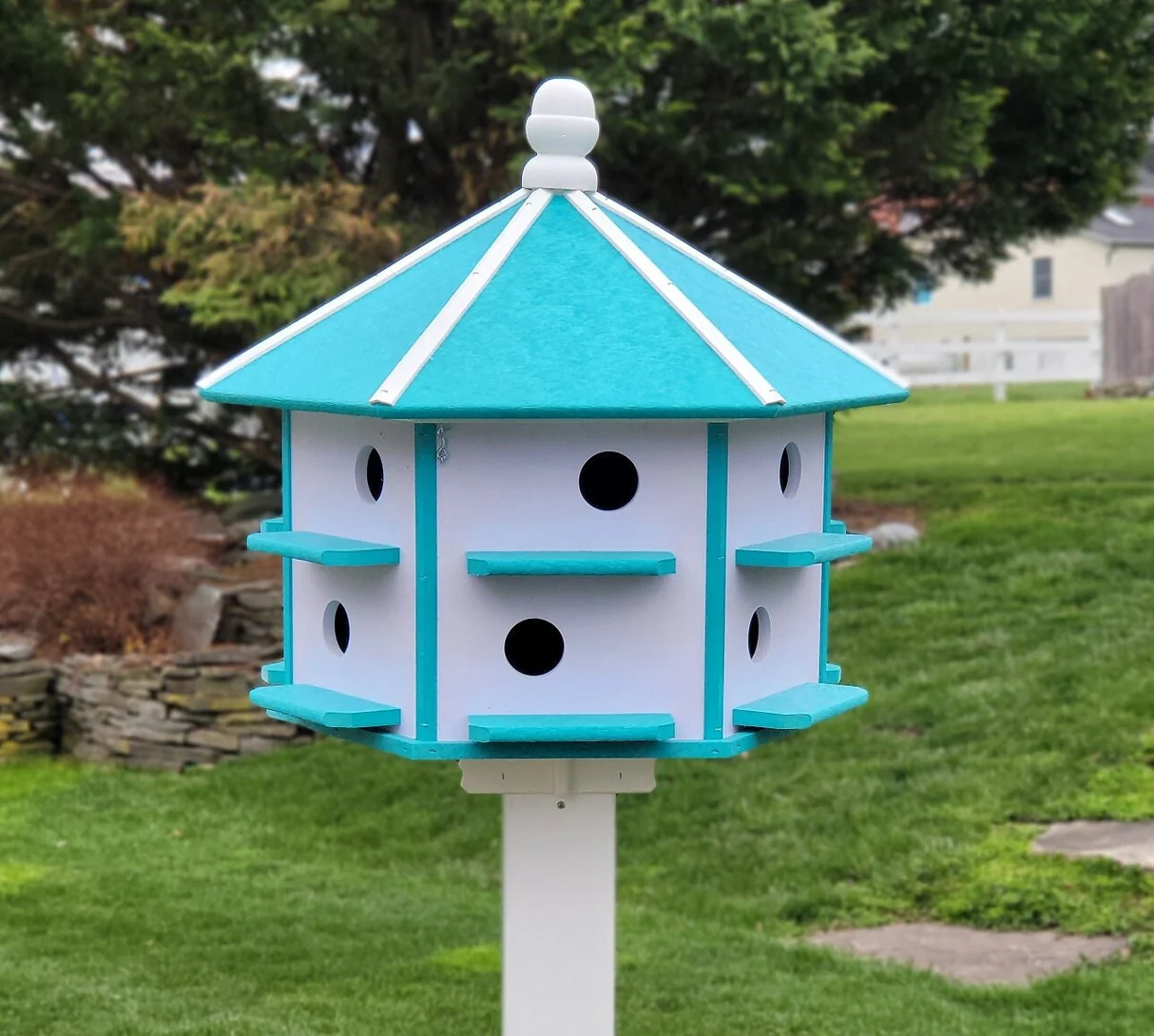 A beautifully crafted Amish-made poly wood Purple Martin birdhouse with 12 nesting compartments, featuring a turquoise and white design, mounted on a post in a lush garden setting.
